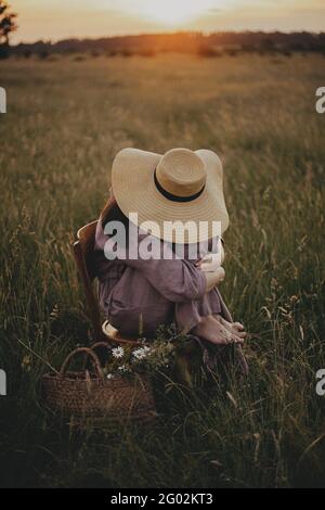 Schöne Frau in Leinenkleid und Strohhut sitzen auf rustikalem Stuhl in Sommerwiese. Junge weibliche Entspannung mit Korb von Blumen in Sonnenuntergang in Coun Stockfoto