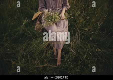 Schöne Frau im Leinenkleid, barfuß auf rustikalem Stuhl sitzend, inmitten von Gras mit Blumenstrauß auf der Sommerwiese. Junge Frau, die sich in Cou entspannt Stockfoto