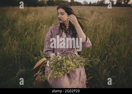 Schöne Frau mit windigen Haaren und in Leinenkleid sitzen auf Holzstuhl mit Blumenstrauß in Sommerwiese. Junge weibliche Entspannung am Abend Stockfoto