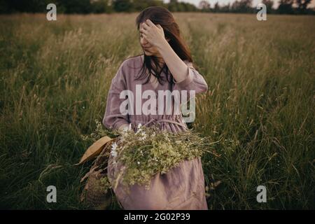 Schöne Frau mit windigen Haaren und in Leinenkleid sitzen auf Holzstuhl mit Blumenstrauß in Sommerwiese. Junge weibliche Entspannung am Abend Stockfoto