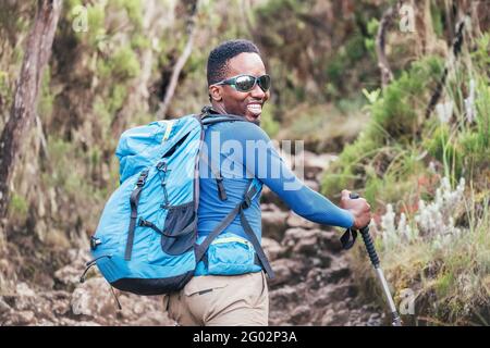 Porträt eines fröhlich lächelnden afroamerikanischen Ethnizität jungen Mannes in Sonnenbrille. Er hat einen Spaziergang mit einem Rucksack mit Trekkingstöcken zu Mweka ga Stockfoto