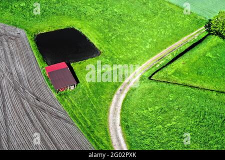 Luftaufnahme eines Schuppen hinter einem kleinen künstlichen Fischteich auf einem Weg durch eine grüne Wiese neben einem Feld, abstrakter Effekt Stockfoto