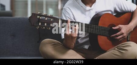 Zugeschnittenes Gelegenheitsfoto, der Gitarre spielt, während er im Wohnzimmer auf dem Sofa sitzt. Stockfoto