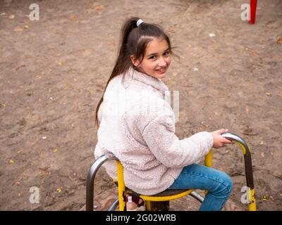 Draufsicht über ein fröhlich lächelndes Teenager-Mädchen (11-12) in einem Kunstpelzmantel und mit langem Pferdeschwanz, der sich bei einer fröhlichen Runde auf dem Spielplatz dreht. Stockfoto