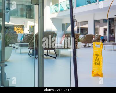 Gelbes Warnschild aus Kunststoff, nasser Fußboden auf der Terrasse eines Hotels in Napa, Zypern. Leere Wicket-Sessel und Sofas mit Kissen im Hintergrund. Stockfoto