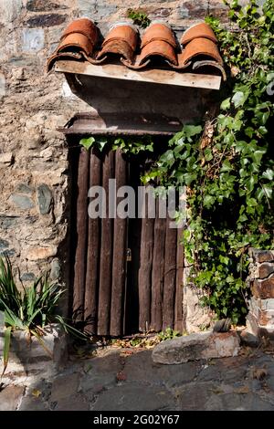 Kleines Fenster eines Steinhauses, von der Sonne beleuchtet, geschlossen mit hölzernen Fensterläden, umgeben von Efeu und geschützt durch ein Ziegeldach. Vertikale Photographer Stockfoto