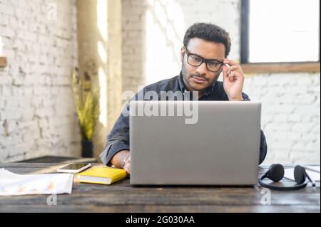 Inteligente indischer Mann mit gemischter Rasse, der auf der Tastatur tippt, Mann in elegantem Casual-Hemd und stilvolle Brillen mit Laptop im modernen Loft-Büro Stockfoto