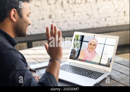 Rückansicht multirassischer Mann aus dem Nahen Osten, der die mobile Computer-App für Videobesprechung mit einer Freundin oder Angestelltin nutzt, die einen Hijab trägt, einen Mann, der einen Laptop benutzt, und online mit einer islamischen Frau spricht. Videoanruf Stockfoto