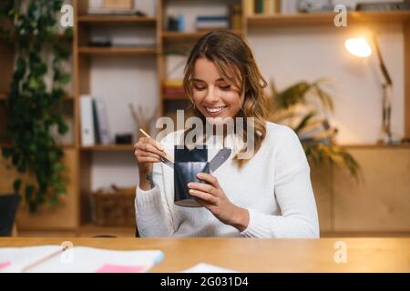 Lächelnde junge Unternehmerin beim Mittagessen, während sie am Schreibtisch sitzt und Nudeln isst Stockfoto