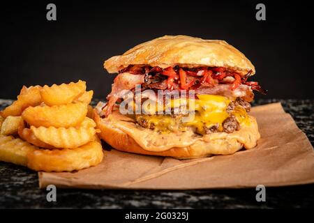 Großer handgemachter Burger mit viel Speck, Käse und Pommes auf schwarzem Hintergrund Stockfoto