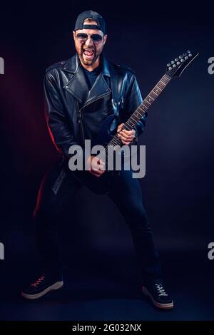 Foto in voller Größe des brutalen bärtigen Heavy Metal-Musikers in Lederjacke und Sonnenbrille spielt sehr emotional elektrische Gitarre. Aufgenommen in einem Stockfoto