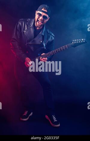 Foto in voller Größe des brutalen bärtigen Heavy Metal-Musikers in Lederjacke und Sonnenbrille spielt sehr emotional elektrische Gitarre. Aufgenommen in einem Stockfoto