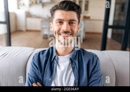 Nahaufnahme eines hübschen, fröhlichen jungen erwachsenen kaukasischen Mannes, der mit gekreuzten Armen auf dem Sofa zu Hause sitzt, in legeres, stilvolles Denim-Hemd gekleidet ist, die Kamera anschaut und freundlich lächelt Stockfoto