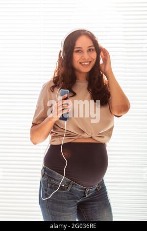 Schöne junge friedliche schwangere Frau hört angenehme klassische Musik mit Smartphone und Kopfhörer. Konzept der positiven Einstellung vor Stockfoto