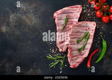 Rohe Schweinefleischrippen oder frisches, ungekochtetes Fleisch mit Gewürzen auf schwarzem Holzblech mit Paprika, Knoblauchzehen und Kräutern. Dunkler strukturierter Hintergrund mit Kopierbereich Stockfoto