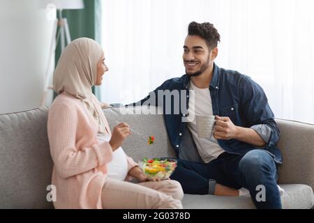 Schwanger Islamische Frau Essen Gemüsesalat, Während Sie Sich Auf Der Couch Ausruhen Mit Ehemann Stockfoto