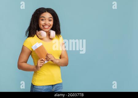 Reisekonzept. Fröhliche afroamerikanische Dame mit Kopfhörern, die Tickets auf blauem Studiohintergrund hält, freier Platz Stockfoto