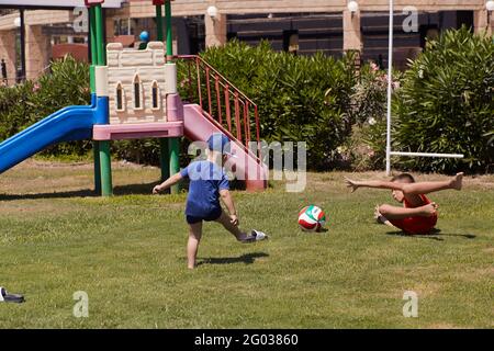 Kemer, Türkei - Mai, 21: Kleinkind und Teenager spielen bei sonnigem Wetter auf grünem, frischem Rasen Fußball. Konzept von sozialer Distanz, Sommerspaß und Urlaub. Hochwertige Fotos Stockfoto