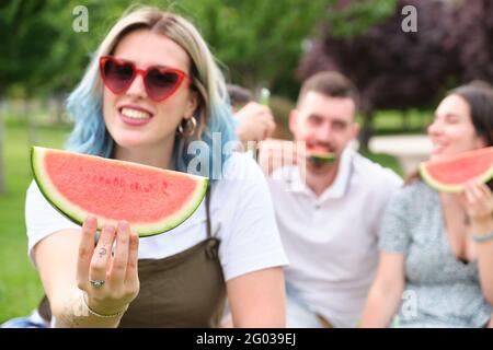 Glückliche junge Frau lächelt, hält ein Stück Wassermelone und zeigt es der Kamera, Freunde essen auf dem Hintergrund. Stockfoto