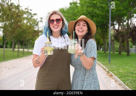 Zwei glückliche Freundinnen trinken erfrischende Getränke und schauen an einem sonnigen Sommertag in einem Park auf die Kamera. Stockfoto