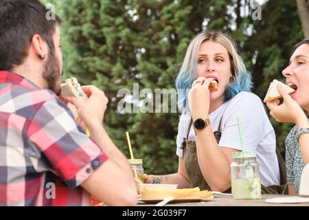 Eine Gruppe von glücklichen Freunden, die Spaß haben, Sandwiches trinken und essen in einem Park. Picknick an einem sonnigen Sommertag. Stockfoto