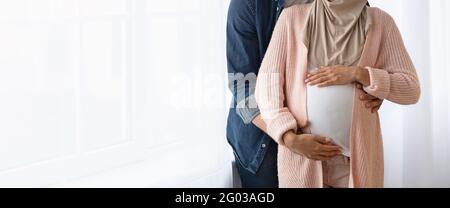 Zugeschnittenes Bild eines muslimischen Paares, das ein Baby erwartet, das sich in der Nähe des Fensters umarmt Zu Hause Stockfoto