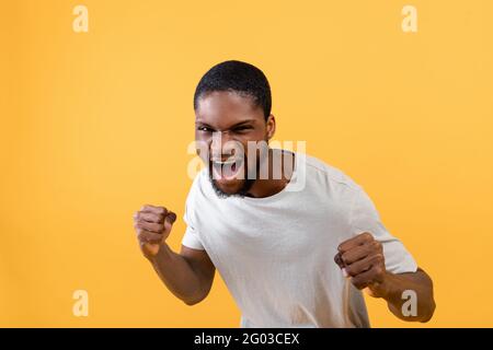Junger schwarzer Mann mit Fäusten-Verteidigungsgeste, schreiend vor Wut, Fäuste auf gelbem Studiohintergrund anklammend Stockfoto
