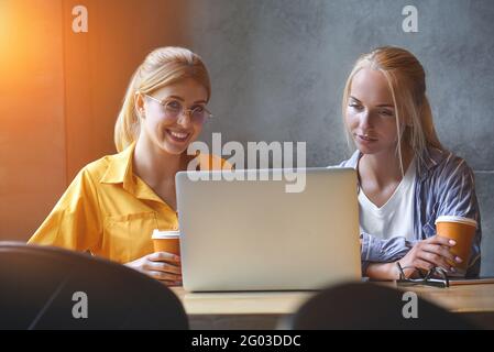 Zwei niedliche Mädchen in legerer Kleidung schauen im Büro auf den Bildschirm eines Laptops. Blogger oder Designer lesen Nachrichten auf einem Laptop Stockfoto