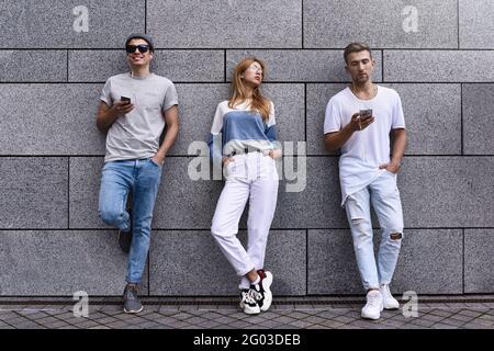 Modetrportrait von drei besten Freunden, die auf der Straße posieren und stilvolles Outfit und Jeans an der grauen Wand tragen Stockfoto