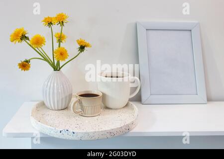 Ein Bouquet von Delelionen in einer weißen, geriffelten Vase und zwei Tassen in verschiedenen Größen mit Kaffee auf einem weißen Tisch und Rahmen für Bilder. Frühstück. SPA kopieren Stockfoto