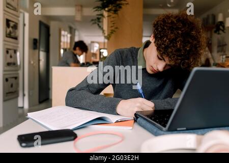 Teenager mit braunem lockigen Haar, der Hausaufgaben macht, während er am Tisch sitzt Stockfoto