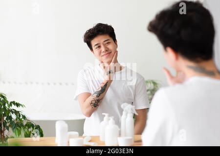 Fröhlicher asiatischer junger Mann mit Tattoos in weißem T-Shirt berührt Wange, genießt sauberes Gesicht Stockfoto