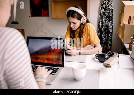 Mädchen mit Kopfhörer E-Learning über digitales Tablet, während sie mit Geschäftsfrau am Schreibtisch sitzt, die am Laptop arbeitet Stockfoto