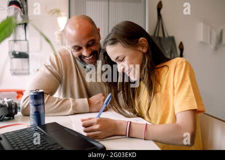 Fröhlicher Vater sitzt mit Tochter Hausaufgaben machen, während am Tisch sitzen Stockfoto