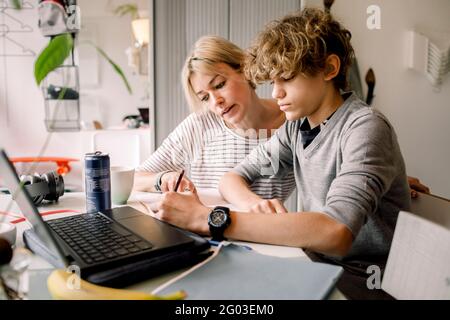 Mutter führt Sohn Hausaufgaben machen, während sie am Tisch sitzt Stockfoto