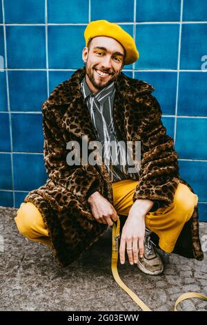 Ein junger Mann mit Mantel, der an der blau gefliesten Wand hocken soll Stockfoto