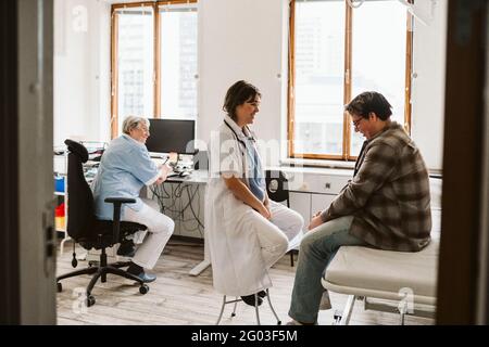 Lächelnde Angestellte im Gesundheitswesen, die mit einem jungen männlichen Patienten spricht, während die ältere Krankenschwester am Schreibtisch sitzt Stockfoto
