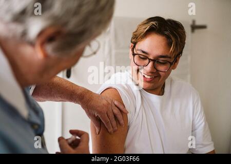 Lächelnder junger männlicher Patient, der eine leitende medizinische Expertin ansieht, die eine Impfung verabreichen kann Stockfoto