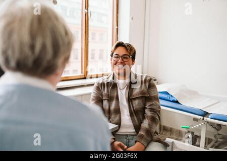 Lächelnder junger männlicher Patient, der in der medizinischen Klinik eine ältere Ärztin ansieht Stockfoto