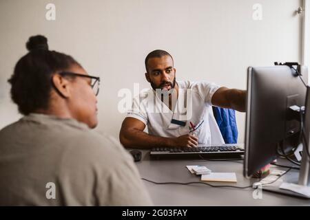 Männlicher Mitarbeiter im Gesundheitswesen, der am Computer mit einer Patientin in einer medizinischen Klinik diskutiert Stockfoto