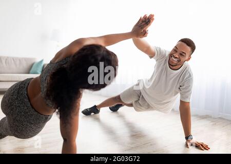 Motiviertes schwarzes Paar, das sich beim Training zu Hause, im Plank stehend und im Team, hohe fünf Punkte gibt Stockfoto