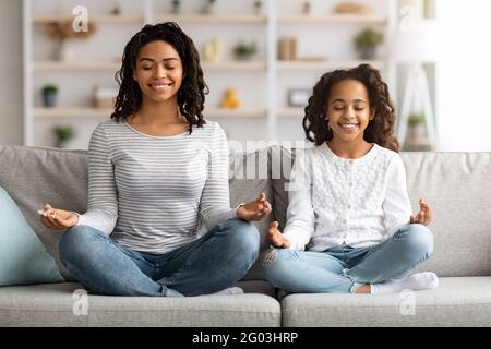 Entspannte schwarze Mutter und Tochter sitzen auf der Couch und meditieren Stockfoto