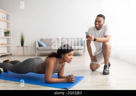 Millennial schwarzes Paar, das zu Hause als Team trainiert, athletische Frau, die in Plankenpose steht und ihr Freund Zeit notiert Stockfoto