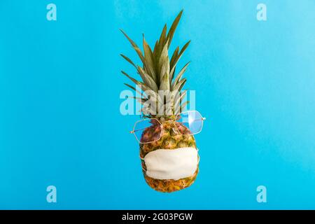 Ananas lustiges Gesicht in stilvollen Sonnenbrillen medizinische Maske auf Farbe blau Sommer Hintergrund. Reisekonzept Ananas in Schutzmaske bei Coronavirus Stockfoto