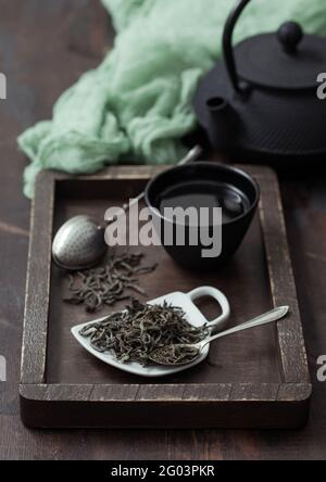 Grüner loser Tee mit Vintage-Sieb-Ei und eiserner Teekane mit Tasse in Holzkiste mit grünem Tuch auf Holzhintergrund. Stockfoto