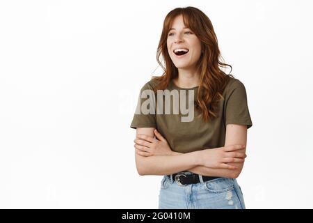 Auffälliges Mädchen, das lachend und mit einem Freund spricht, die Arme auf der Brust kreuzt, die Person beiseite schaut, etwas Lustiges beobachtet, im T-Shirt gegen Weiß stehend Stockfoto