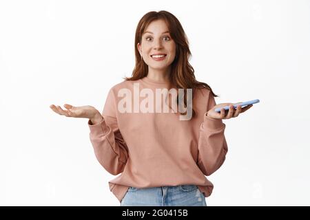 Lächelnd überrascht Rotschopf Mädchen, hält Smartphone und zuckende Schultern, erhalten erstaunliche Nachrichten auf Handy, stehend über weißem Hintergrund Stockfoto