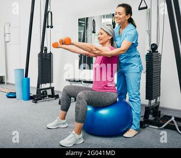 Physiotherapeutin, die mit reifen weiblichen Patienten in einem Reha-Zentrum arbeitet. Eine Chinesin hebt mit einem Personal Trainer Hanteln auf einem Fitnessball Stockfoto