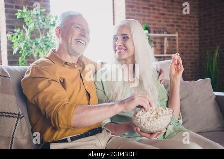 Foto von fröhlich grauhaarigen Paar sitzen Couch essen Pop Mais sehen tv Kino Lächeln drinnen im Haus Wohnung Stockfoto