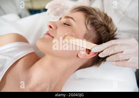 Nahaufnahme der Hand der Kosmetikerin mit Holzspachtel zum Auftragen transparent Anbestimmt-Gel auf dem Gesicht der Frau für die Laserentfernung Haarverfahren n Stockfoto
